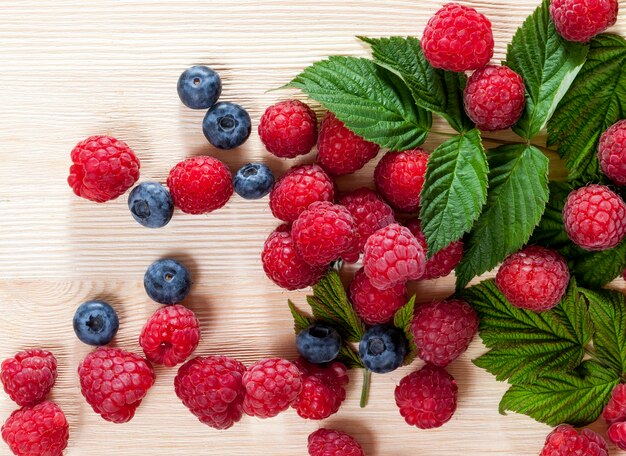 , framboises mûres et myrtilles juteuses sur une planche de bois, les framboises rouges sont couvertes de feuilles feuillues, gros plan en été