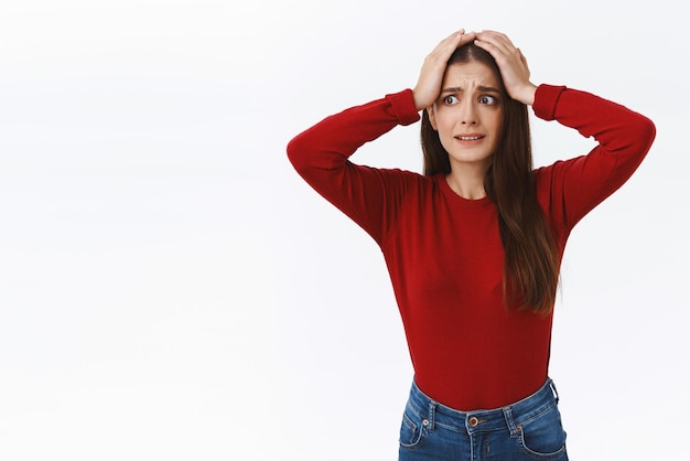 Freakout alarmé jeune fille brune gênante en pull rouge attrape la tête et regarde dans la panique debout inquiet ou désespéré debout dérangé préoccupé par la situation choquante avoir l'air effrayé