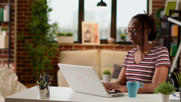 Freelancer afro-américain tapant des informations sur un ordinateur portable, travaillant à distance depuis son bureau à domicile. Jeune femme naviguant sur Internet sur ordinateur pour effectuer un travail à distance, un travail commercial en ligne distant. Prise de vue au trépied.