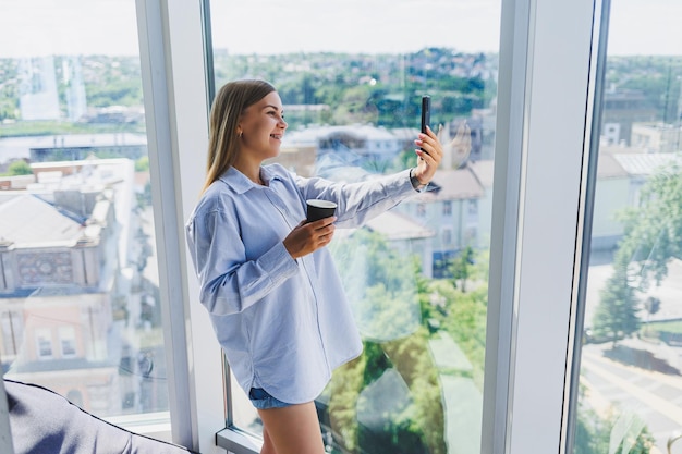 Freelancer de femme moderne parlant au téléphone tout en se tenant devant une grande fenêtre et en buvant du café Le concept d'une femme moderne réussie Jeune fille dans un bureau ouvert