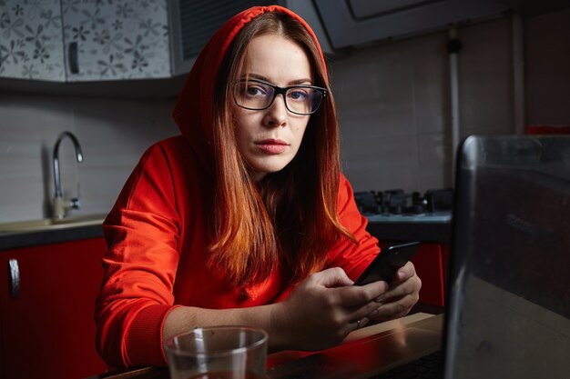 Freelancer vêtu d'un sweat-shirt rouge à l'aide de téléphone et de boire du café dans la cuisine