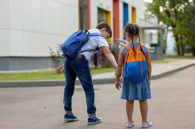 Un frère d'écolier avec un sac à dos bleu prend sa petite sœur par la main pour entrer dans le