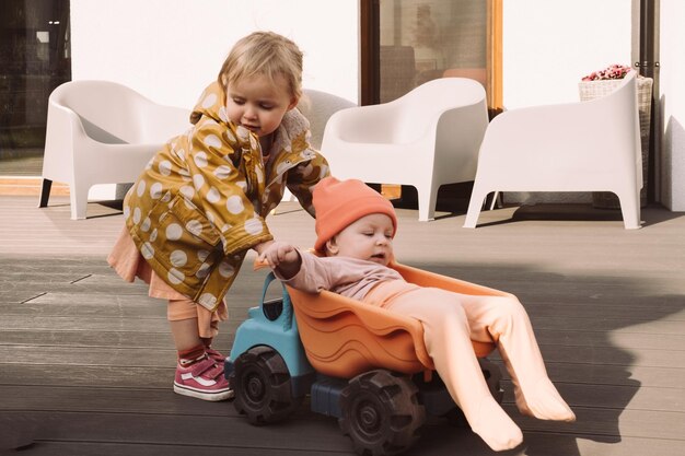 Photo un frère et une sœur conduisent leur petite sœur dans une grosse voiture de jouet à l'extérieur. des enfants s'amusent près de la maison.