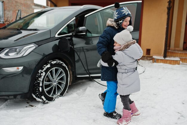 Frère et soeur jouent contre la charge d'une voiture électrique dans la cour de la maison en hiver