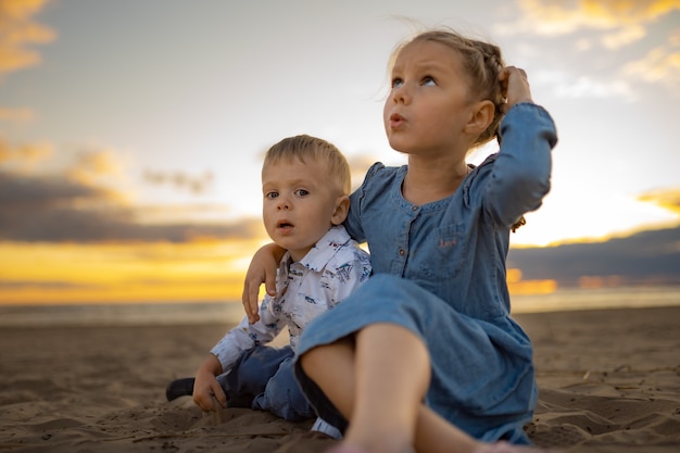 frère et soeur mignons assis sur le sable à la plage au coucher du soleil