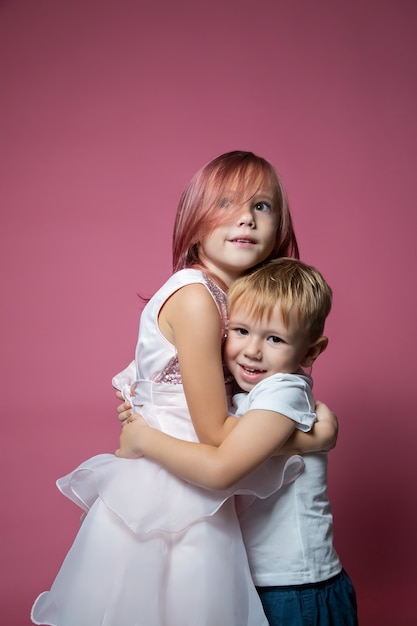 Frère et sœur de race blanche, étreignant la caméra sur fond rose tourné en studio. Liens familiaux, amitié, concept d'enfance heureuse.