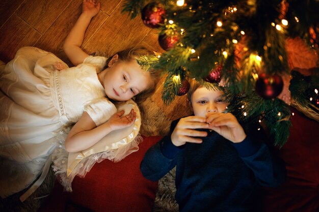Frère et sœur se trouvent sur le tapis près de l'arbre du Nouvel An dans un intérieur de Noël confortable