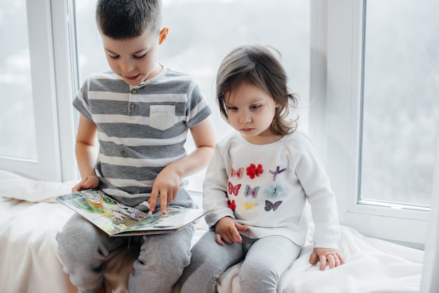 Frère et sœur sont assis sur le rebord de la fenêtre et lisent un livre. Bonheur, famille.