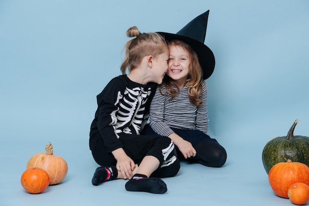 Frères et sœurs en costumes d'halloween touchant des têtes et riant. Assis par terre entre de petites citrouilles. Sur fond bleu.