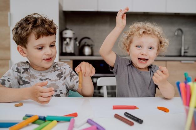 Les frères et sœurs heureux sont assis à la table de la cuisine et jouent avec de la pâte à modeler un garçon d'âge scolaire