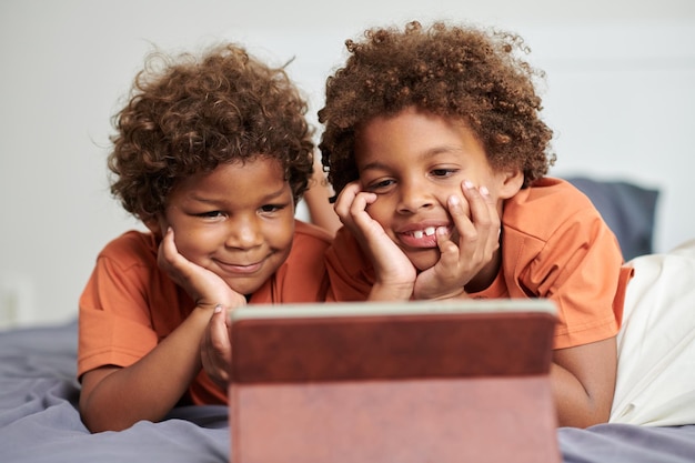 Des frères et sœurs joyeux allongés sur le lit et regardant des films pour enfants sur leur tablette.