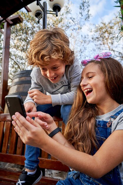 Des frères et sœurs joyeux en tenue décontractée souriant et regardant des vidéos sur un téléphone portable tout en étant assis ensemble sur un banc en bois dans le parc le jour d'été