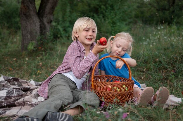 Frères et sœurs souriants avec des pommes à la main s'asseoir sur la pelouse sur le couvre-lit dans le jardin Pique-nique pour les enfants lors d'une chaude journée d'été