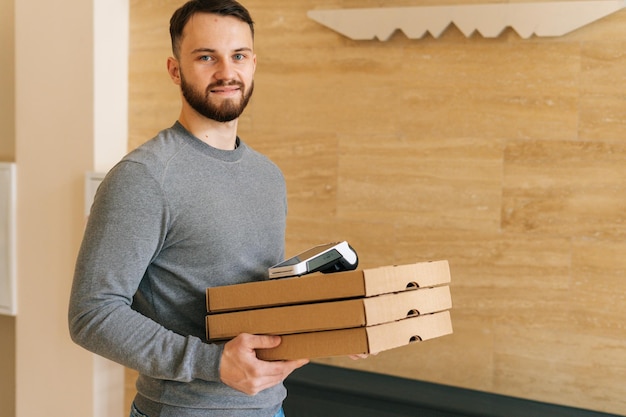Friedly courier male holding boxes pizza et terminal de paiement sans contact debout dans le hall d'entrée d'un appartement ou d'un immeuble de bureaux