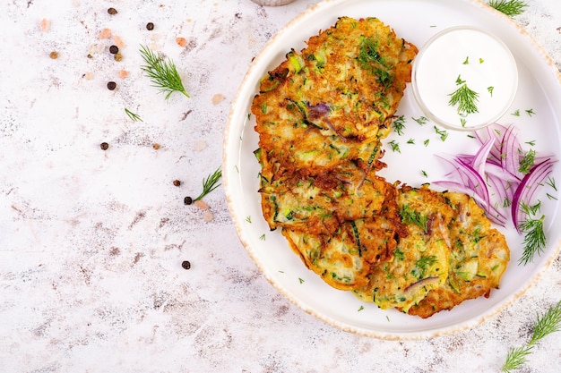 Frites de courgette avec oignons rouges, ail et herbes, crêpes végétariennes de courgette et crème sure sur table blanche.