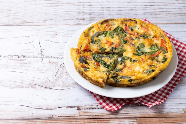 Photo frittata italienne faite avec des épinards, des tomates, des oignons et des poivrons sur une table en bois blanc