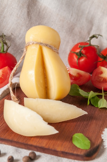 Fromage Caciocavallo sur une planche de bois rectangulaire. Deux morceaux en sont coupés. Décoré avec de la roquette, des épices et des tomates. Tissu de lin gris de fond. Fermer.