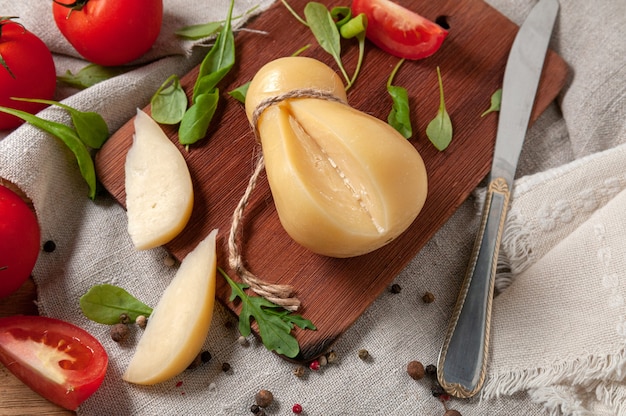 Le fromage Caciocavallo repose sur une planche de bois rectangulaire. Deux morceaux sont coupés. Décoré avec de la roquette, des épices et des tomates. Tissu de lin gris de fond. Vue d'en-haut.