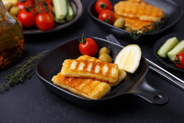 fromage halloumi grillé dans une casserole noire avec des olives, des tomates, des concombres, du thym et du citron fromage traditionnel de la Méditerranée orientale à fond noir en gros plan sélectif