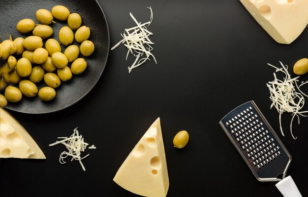 Fromage et olives sur fond noir. Composition à plat avec des morceaux de fromage, une râpe et un plat aux olives.