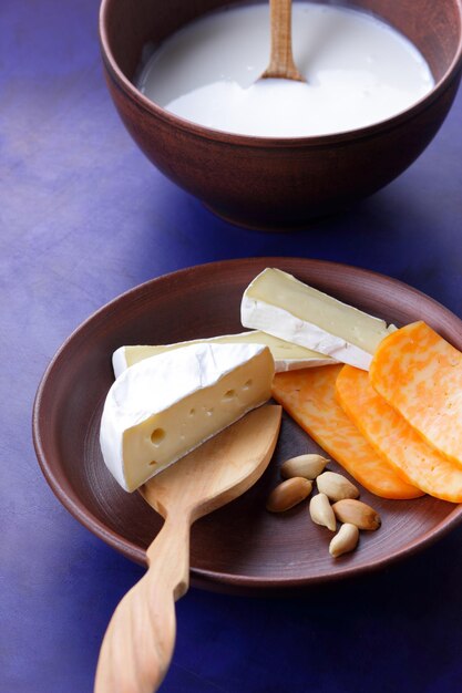 Fromages aux noix et une spatule en bois sur une plaque d'argile Différents types d'assiette de fromage et un bol de lait sur un gros plan de fond bleu