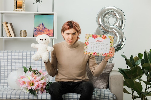 Fronçant les sourcils beau mec le jour de la femme heureuse tenant un ours en peluche avec un calendrier assis sur un canapé dans le salon