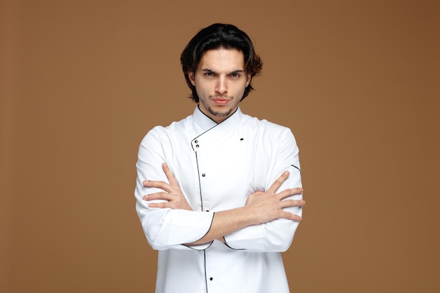 fronçant les sourcils jeune chef masculin portant l'uniforme regardant la caméra tout en gardant les bras croisés isolé sur fond marron