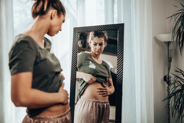 Frown jeune femme debout devant un miroir et se tenant la main sur son ventre gonflé.