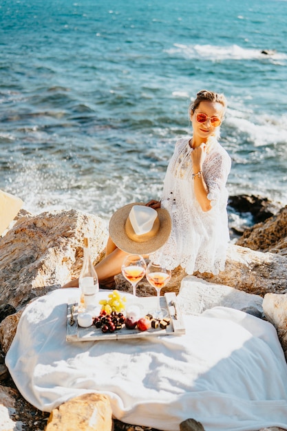 Fruits, collations et boissons pendant les pique-niques en mer. Fille faisant des farcis au bord de la mer avec des rochers, vêtue d'une élégante robe blanche, chapeau de paille. Concept de voyage d'agriculture de tourisme. Voyage dans les pays du sud