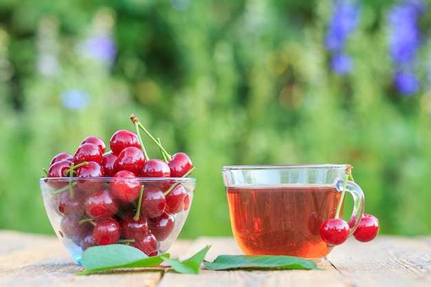 Fruits rouges de cerise avec des pédoncules dans un bol en verre et une tasse de thé sur de vieilles planches en bois.