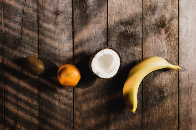 Fruits tropicaux: noix de coco cassée, kiwi, mandarine, orange, banane sur fond en bois marron avec ombre de feuille de palmier. Pulpe de noix de coco blanche. Photo de haute qualité