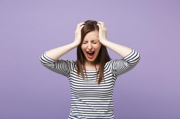 Frustré pleurer jeune femme brune fille dans des vêtements rayés décontractés posant isolé sur fond violet violet portrait en studio. Concept de style de vie des gens. Maquette de l'espace de copie. Mettre les mains sur la tête.