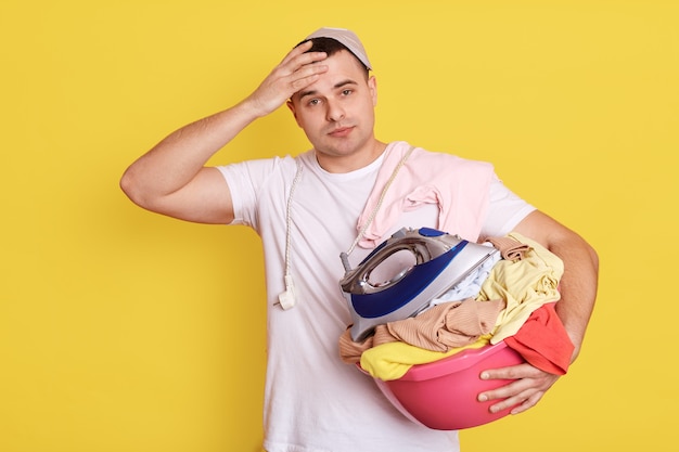 Frustré, surmené, homme posant avec bassin plein de vêtements sales, ayant beaucoup de tâches à la maison, se tient avec un panier à linge, homme portant un t-shirt blanc isolé sur un mur jaune.