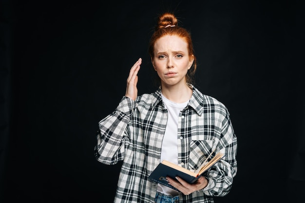 Frustrée bouleversée jeune femme étudiante tenant un livre et regardant la caméra sur fond noir isolé Joli modèle de dame rousse montrant émotionnellement des expressions faciales dans l'espace de copie de studio