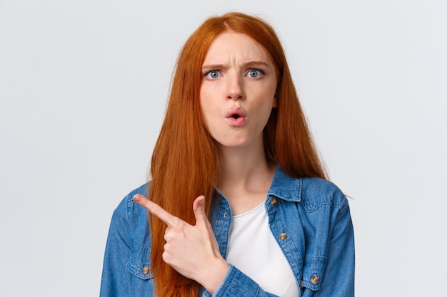 Frustrée et folle, femme rousse se plaignant en colère se disputant avec son frère, pointant le coin supérieur gauche ou derrière, pliant les lèvres et fronçant les sourcils, debout perplexe sur le mur blanc