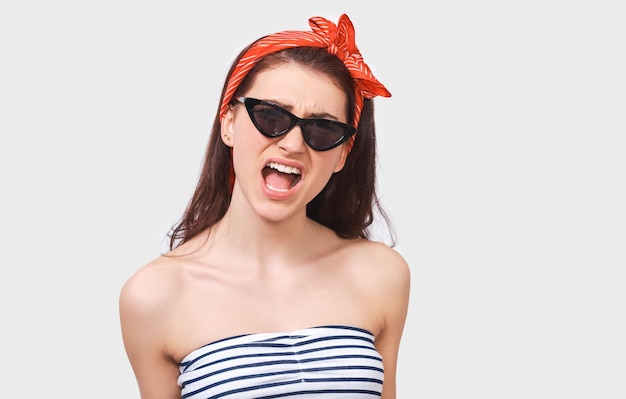 Frustrée jeune femme brune ouvre la bouche largement crie fort porte des lunettes de soleil noires à la mode bandeau rouge et chemisier rayé posant contre un mur blanc Étudiante nerveuse criant