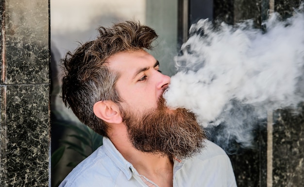 Fumer la cigarette électronique. Concept de soulagement du stress. Appareil à fumer. Homme longue barbe détendue avec l'habitude de fumer. Nuages de fumée aromatisée. Homme barbu fumant de la vape. L'homme à la barbe expire de la fumée.