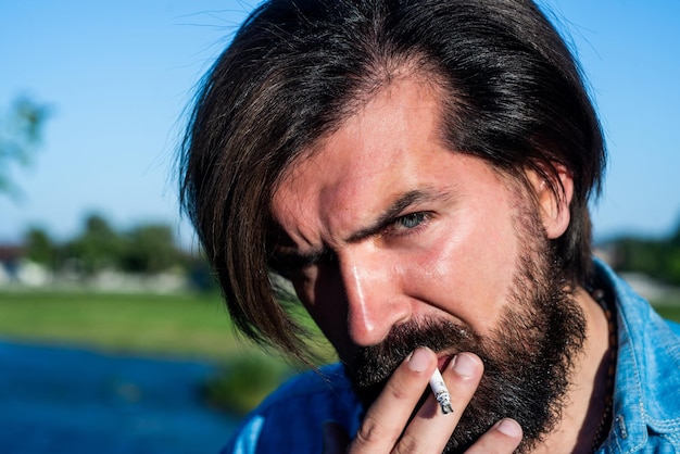 Fumeur brutal hipster caucasien brutal avec moustache fumant une cigarette confiant beau mec portrait de printemps homme barbu brutal porter une chemise décontractée et des lunettes de soleil mode et beauté d'été masculin