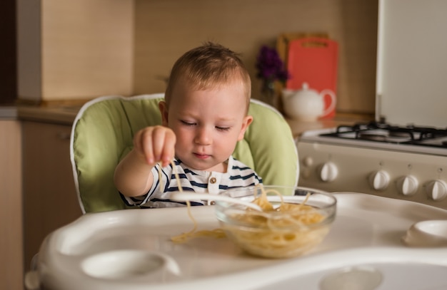 Funny boy mange des spaghettis avec ses mains. Un petit garçon est assis sur une chaise haute dans la cuisine.