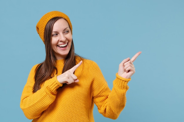 Funny young brunette woman girl in yellow pull hat posant isolé sur fond bleu studio portrait. Concept de mode de vie des émotions sincères des gens. Maquette de l'espace de copie. Pointer les index de côté.