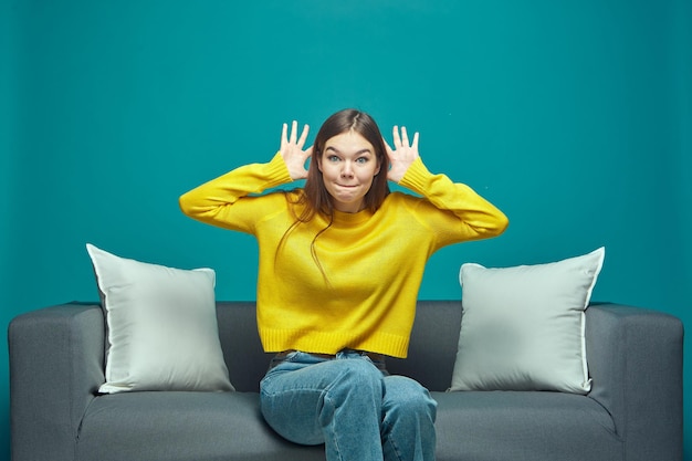 Funny young girl making faces grimaçant tromper montrant de grandes oreilles par les mains taquiner sitting on sofa