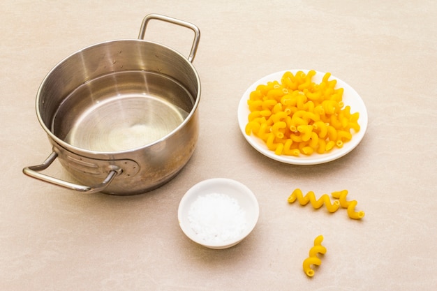 Fuzilli cru avec de l'eau en pot et du sel. Pâtes italiennes traditionnelles en plaque blanche sur une surface en pierre. Préparation pour les enfants, gros plan