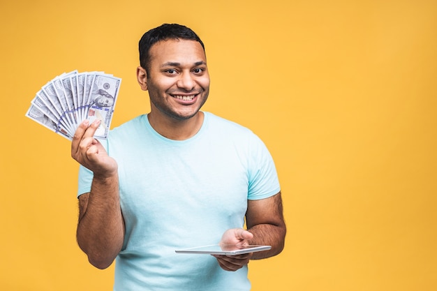 Gagnant! Jeune homme noir afro-américain heureux riche en tenue décontractée de billets d'un dollar avec surprise isolée sur fond de mur jaune. Utilisation de la tablette.