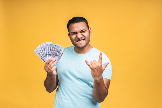 Gagnant! Jeune homme noir afro-américain heureux riche en tenue décontractée de billets d'un dollar avec surprise isolée sur fond de mur jaune.