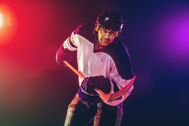 Gagnant. Joueur de hockey masculin avec le bâton sur la glace et le mur de couleur néon foncé. Sportif portant des équipements, casque pratiquant. Concept de sport, mode de vie sain, mouvement, bien-être, action.