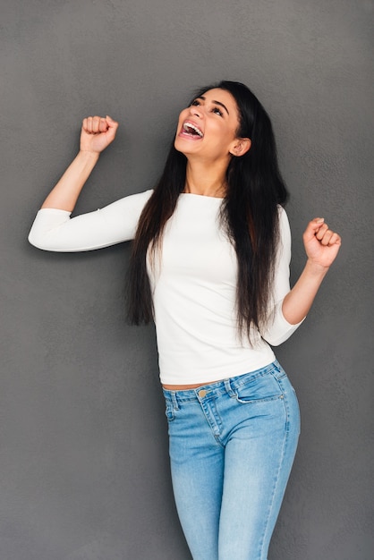 Gagnant de tous les jours. Jeune femme africaine excitée gardant les bras levés et exprimant sa positivité en se tenant debout sur fond gris