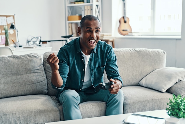 Gagner le match. Beau jeune homme africain faisant des gestes et souriant alors qu'il était assis à l'intérieur