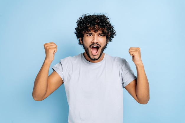 Gagnez le triomphe Un indien ou un arabe étonné excité en t-shirt se réjouit de la victoire de la victoire en gesticulant avec les poings en regardant la caméra en souriant debout sur un fond bleu isolé en criant