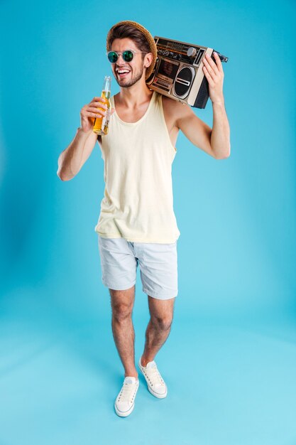 Gai jeune homme au chapeau et lunettes de soleil avec boombox marchant et buvant de la bière sur le mur bleu