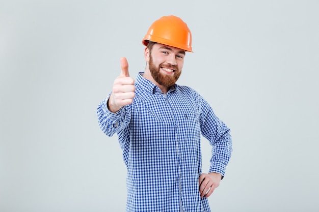 Gai jeune homme barbu en casque debout et montrant les pouces vers le haut sur le mur blanc